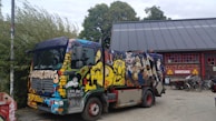A colorfully painted garbage truck is parked next to a building with a large rooftop. The truck is adorned with vibrant graffiti art featuring various characters and designs. Nearby, there are bicycles and a sign that says 'Christiania' on the building, suggesting a creative and alternative atmosphere. Trees and bushes surround the area, adding a natural backdrop to the urban setting.