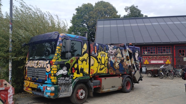 A colorfully painted garbage truck is parked next to a building with a large rooftop. The truck is adorned with vibrant graffiti art featuring various characters and designs. Nearby, there are bicycles and a sign that says 'Christiania' on the building, suggesting a creative and alternative atmosphere. Trees and bushes surround the area, adding a natural backdrop to the urban setting.