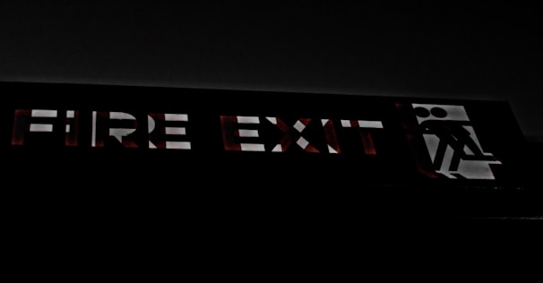 A dimly lit fire exit sign with bold, uppercase letters glowing faintly against a dark background. The sign includes a pictogram of a person walking through a doorway, indicating the exit route.