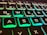 Close-up view of an illuminated computer keyboard featuring keys highlighted with a green and blue backlight. The focus is mainly on the QWERTY layout, with keys such as Q, W, E, A, S, and D clearly visible.