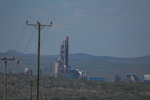 A large industrial facility or factory with tall, cylindrical structures and various piping against a backdrop of rolling hills. There are power lines in the foreground stretching across the landscape.