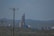 A large industrial facility or factory with tall, cylindrical structures and various piping against a backdrop of rolling hills. There are power lines in the foreground stretching across the landscape.