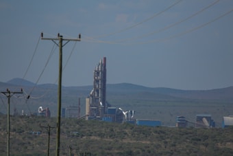 A large industrial facility or factory with tall, cylindrical structures and various piping against a backdrop of rolling hills. There are power lines in the foreground stretching across the landscape.