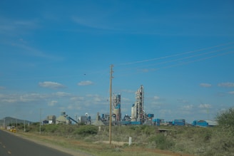 A large industrial complex with several tall buildings and towers stands in a rural setting. Power lines stretch across the sky, which is clear with scattered clouds. Greenery and bushes surround the factory area with an open road leading towards it.