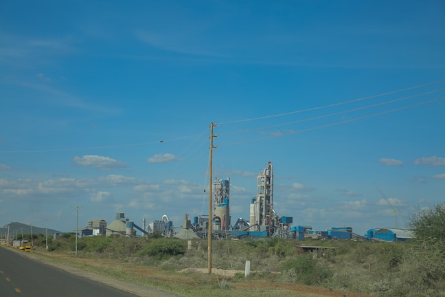 A large industrial complex with several tall buildings and towers stands in a rural setting. Power lines stretch across the sky, which is clear with scattered clouds. Greenery and bushes surround the factory area with an open road leading towards it.