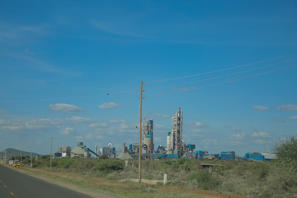 A large industrial complex with several tall buildings and towers stands in a rural setting. Power lines stretch across the sky, which is clear with scattered clouds. Greenery and bushes surround the factory area with an open road leading towards it.