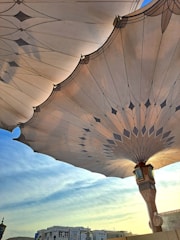 Large architectural umbrellas with intricate designs stretch across the sky, partially shading buildings below. The patterning on the umbrellas features geometric shapes and is highlighted by the sun's rays. In the background, a clear blue sky transitions into a softer hue near the horizon, while the buildings display a blend of modern and traditional architectural elements.