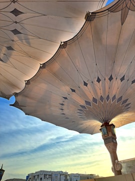 Large architectural umbrellas with intricate designs stretch across the sky, partially shading buildings below. The patterning on the umbrellas features geometric shapes and is highlighted by the sun's rays. In the background, a clear blue sky transitions into a softer hue near the horizon, while the buildings display a blend of modern and traditional architectural elements.