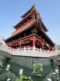 A traditional multi-tiered pagoda with vibrant red and gold colors, decorative roof elements, and a surrounding stone railing. The structure is reflected in a pond below, which contains floating lotus flowers and lanterns, enhancing the serene ambiance.