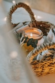 A lit candle inside a glass jar is decorated with eucalyptus leaves and an orange slice. It is placed in a woven basket along with white towels, creating a cozy and warm atmosphere.