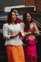 Two women dressed in elegant traditional clothing stand side by side, holding their hands in a gesture of respect or prayer. A person with a painted face in a mask stands behind them, partially concealed. The women are in front of a black car, with an urban backdrop featuring a brick wall and some small building structures.