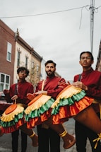 Three individuals dressed in red and maroon outfits are holding large percussion instruments with colorful, fringed covers featuring yellow, green, and orange hues. They are standing on a street lined with brick buildings. The sky is overcast, adding a subdued atmosphere to the urban setting.