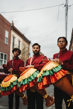 Three individuals dressed in red and maroon outfits are holding large percussion instruments with colorful, fringed covers featuring yellow, green, and orange hues. They are standing on a street lined with brick buildings. The sky is overcast, adding a subdued atmosphere to the urban setting.