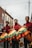 Three individuals dressed in red and maroon outfits are holding large percussion instruments with colorful, fringed covers featuring yellow, green, and orange hues. They are standing on a street lined with brick buildings. The sky is overcast, adding a subdued atmosphere to the urban setting.