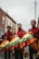 Three individuals dressed in red and maroon outfits are holding large percussion instruments with colorful, fringed covers featuring yellow, green, and orange hues. They are standing on a street lined with brick buildings. The sky is overcast, adding a subdued atmosphere to the urban setting.