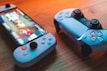 A gaming scenario featuring a smartphone with a game interface next to two controllers on a wooden surface. The smartphone displays vibrant images and icons, while the controllers feature a sleek, modern design with buttons and joysticks prominently visible. Warm lighting enhances the scene.