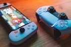 A gaming scenario featuring a smartphone with a game interface next to two controllers on a wooden surface. The smartphone displays vibrant images and icons, while the controllers feature a sleek, modern design with buttons and joysticks prominently visible. Warm lighting enhances the scene.