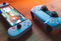 A gaming scenario featuring a smartphone with a game interface next to two controllers on a wooden surface. The smartphone displays vibrant images and icons, while the controllers feature a sleek, modern design with buttons and joysticks prominently visible. Warm lighting enhances the scene.