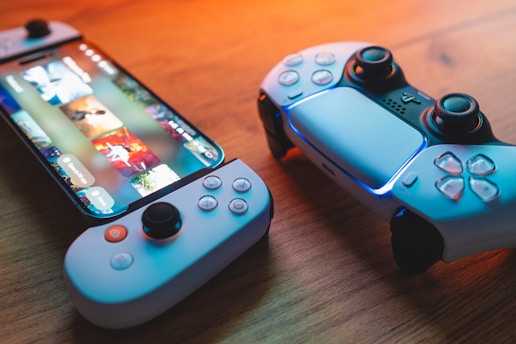 A gaming scenario featuring a smartphone with a game interface next to two controllers on a wooden surface. The smartphone displays vibrant images and icons, while the controllers feature a sleek, modern design with buttons and joysticks prominently visible. Warm lighting enhances the scene.