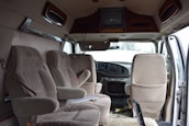 The interior of a van is shown with three beige upholstered seats arranged in two rows. The dashboard is visible, featuring a steering wheel and controls. A television is mounted on the ceiling, and papers are scattered on the floor. The door on the right is open, allowing light to illuminate the cabin.