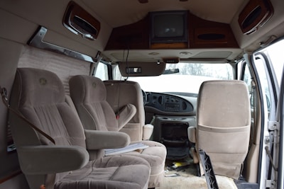 The interior of a van is shown with three beige upholstered seats arranged in two rows. The dashboard is visible, featuring a steering wheel and controls. A television is mounted on the ceiling, and papers are scattered on the floor. The door on the right is open, allowing light to illuminate the cabin.
