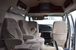 The interior of a van is shown with three beige upholstered seats arranged in two rows. The dashboard is visible, featuring a steering wheel and controls. A television is mounted on the ceiling, and papers are scattered on the floor. The door on the right is open, allowing light to illuminate the cabin.