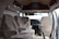 The interior of a van is shown with three beige upholstered seats arranged in two rows. The dashboard is visible, featuring a steering wheel and controls. A television is mounted on the ceiling, and papers are scattered on the floor. The door on the right is open, allowing light to illuminate the cabin.