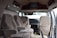 The interior of a van is shown with three beige upholstered seats arranged in two rows. The dashboard is visible, featuring a steering wheel and controls. A television is mounted on the ceiling, and papers are scattered on the floor. The door on the right is open, allowing light to illuminate the cabin.