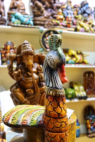 A variety of colorful wooden sculptures are displayed on shelves. The foreground features a brightly painted peacock sculpture with intricate patterns. Behind it, a detailed statue of the Hindu deity Ganesha can be seen, adorned with traditional embellishments. The background is filled with various other handcrafted figures, each with different colors and detailing.