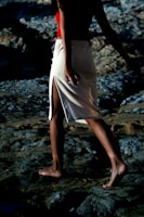 A person walks barefoot on rocky terrain, wearing a red top and a beige skirt with a side slit. The lighting creates deep shadows, adding contrast to the rough texture of the rocks.