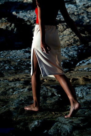 A person walks barefoot on rocky terrain, wearing a red top and a beige skirt with a side slit. The lighting creates deep shadows, adding contrast to the rough texture of the rocks.