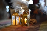 Two decorative lanterns with lit candles inside are placed on a table. The lanterns are ornate with intricate designs, and they create a warm and cozy ambience. Behind the lanterns, there is a glass jar with lights inside and some blurred decorative flowers.