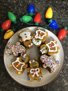 A plate of gingerbread cookies decorated with colorful icing and sprinkles is placed on a dark countertop next to a string of large, colorful Christmas lights. The cookies are shaped like gingerbread people, a house, and other festive shapes. Each cookie is uniquely decorated with various patterns and colors, including white icing and multicolored candy pieces.