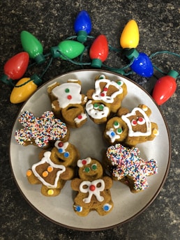 A plate of gingerbread cookies decorated with colorful icing and sprinkles is placed on a dark countertop next to a string of large, colorful Christmas lights. The cookies are shaped like gingerbread people, a house, and other festive shapes. Each cookie is uniquely decorated with various patterns and colors, including white icing and multicolored candy pieces.