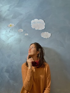 A person is looking thoughtfully upwards while touching their chin. They are standing against a textured blue wall with cut-out paper clouds that have words written on them, including 'progress' and 'in'. The person is wearing glasses and a brown sweater.