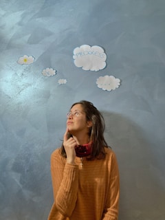A person is looking thoughtfully upwards while touching their chin. They are standing against a textured blue wall with cut-out paper clouds that have words written on them, including 'progress' and 'in'. The person is wearing glasses and a brown sweater.
