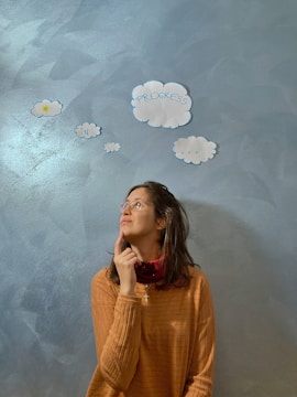 A person is looking thoughtfully upwards while touching their chin. They are standing against a textured blue wall with cut-out paper clouds that have words written on them, including 'progress' and 'in'. The person is wearing glasses and a brown sweater.