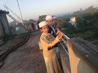 Three children are standing on a rooftop, leaning against a railing and looking into the distance. They are wearing traditional attire and white caps. The background features trees, buildings, and a clear sky, with another group of people visible further back.