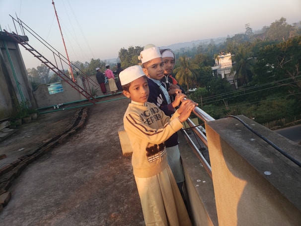 Three children are standing on a rooftop, leaning against a railing and looking into the distance. They are wearing traditional attire and white caps. The background features trees, buildings, and a clear sky, with another group of people visible further back.