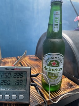 A Heineken Silver beer bottle stands on a wooden surface next to a digital thermometer. The thermometer displays several temperature readings. The background is a soft blue color, enhancing the refreshing theme.