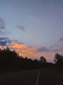 A winding road surrounded by dark silhouettes of trees under a sky with soft orange and blue hues. The clouds are dispersed, with the fading sunlight casting a glow in the horizon.