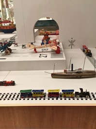 A display of vintage toys including a colorful wooden train set on tracks, multiple model airplanes, a toy ship, and other small toy vehicles arranged in a diorama-like setting. The background features signage and architectural elements that resemble a transportation or airport theme.