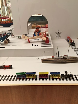 A display of vintage toys including a colorful wooden train set on tracks, multiple model airplanes, a toy ship, and other small toy vehicles arranged in a diorama-like setting. The background features signage and architectural elements that resemble a transportation or airport theme.