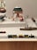 A display of vintage toys including a colorful wooden train set on tracks, multiple model airplanes, a toy ship, and other small toy vehicles arranged in a diorama-like setting. The background features signage and architectural elements that resemble a transportation or airport theme.