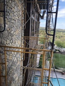 A construction site featuring scaffolding along a stone-finished building wall. The structure appears to be several stories high, with scaffolding made of rusty yellow metal. Beyond the scaffolding, there is a view of a pool, neatly trimmed lawns, and a distant landscape with trees and a road. The sky is partly cloudy, adding a natural backdrop to the scene.