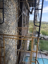 A construction site featuring scaffolding along a stone-finished building wall. The structure appears to be several stories high, with scaffolding made of rusty yellow metal. Beyond the scaffolding, there is a view of a pool, neatly trimmed lawns, and a distant landscape with trees and a road. The sky is partly cloudy, adding a natural backdrop to the scene.