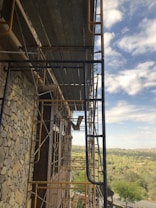 A construction site with metal scaffolding attached to the side of a building. The building has a stone facade and large windows. The scene includes a view of the countryside, with a mix of trees and open land extending into the distance under a partly cloudy sky.