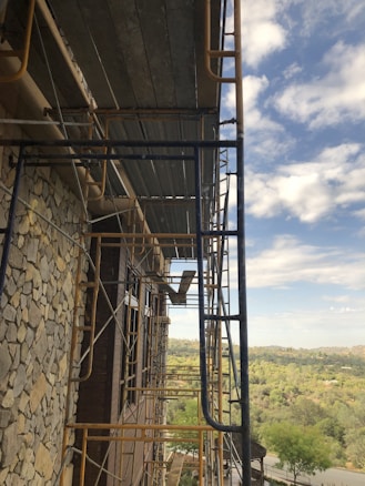 A construction site with metal scaffolding attached to the side of a building. The building has a stone facade and large windows. The scene includes a view of the countryside, with a mix of trees and open land extending into the distance under a partly cloudy sky.