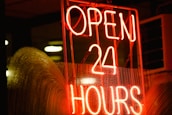 A bright neon sign with the words 'OPEN 24 HOURS' in bold red letters. The sign is lit up against a dark background, with droplets of water visible on the glass surface in front of the sign, likely due to rain.