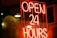 A bright neon sign with the words 'OPEN 24 HOURS' in bold red letters. The sign is lit up against a dark background, with droplets of water visible on the glass surface in front of the sign, likely due to rain.